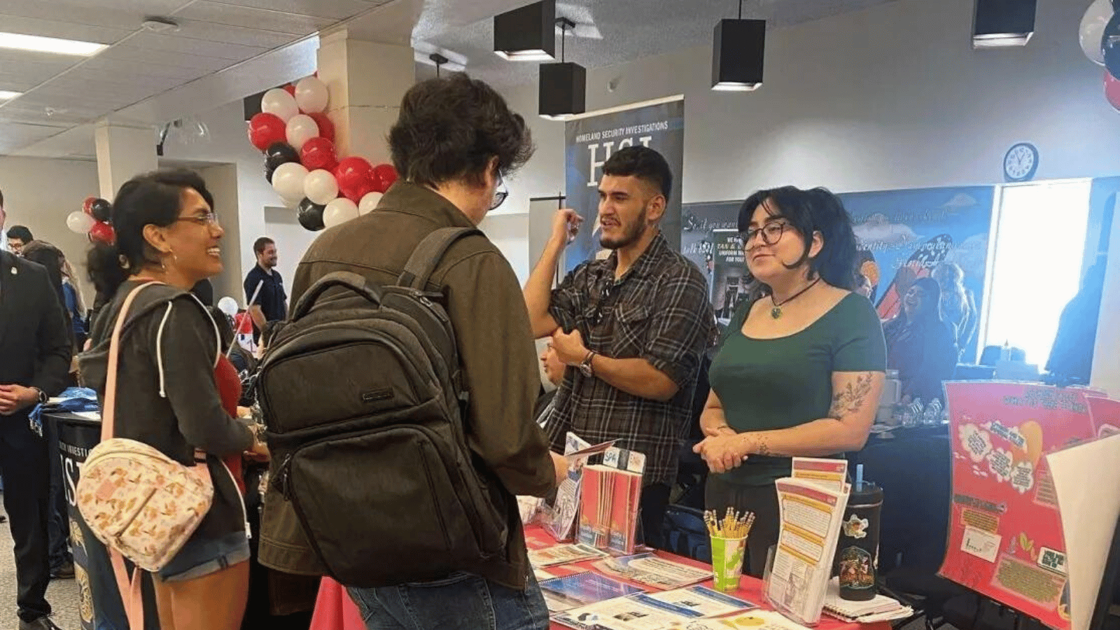 Two recruiters speaking to two students at a career fair booth. On the table between them there are flyers, brochures and pens.